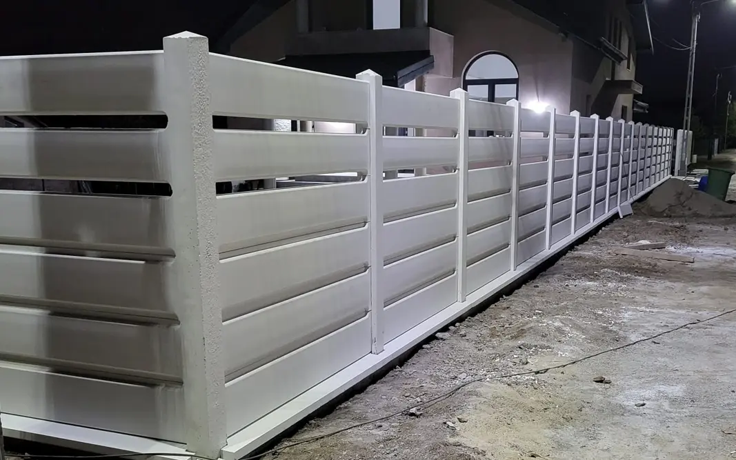 Bílý betonový plot s vodorovnými deskami před domem, vyfocený večer