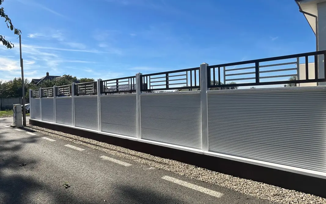 Bílý betonový plot s kovovou mříží nad deskami, u ulice